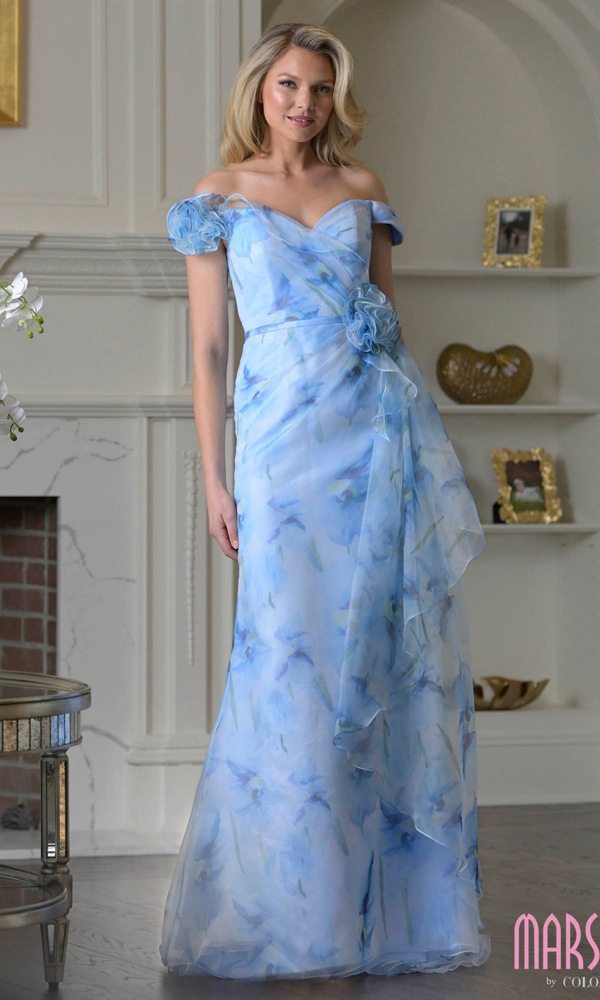 A woman wearing the blue multi Marsoni MV1394 long formal dress with off-the-shoulder floral details and a ruffled flower at the waist stands in a living room with white walls and shelves.